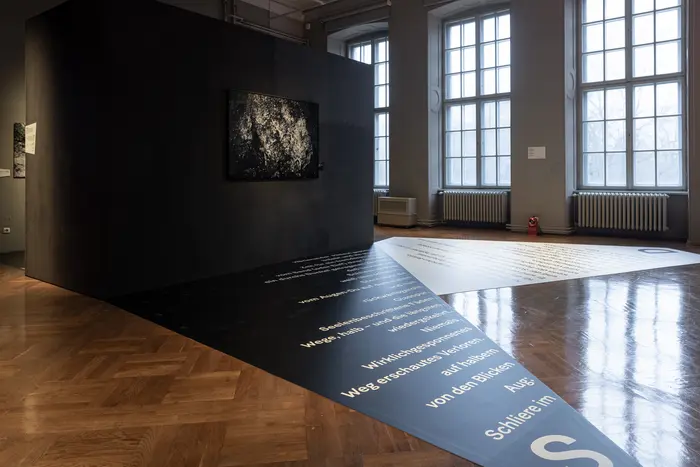 Ein Raum mit Fenstern und einer dunklen Wand, an welcher ein Gemälde hängt