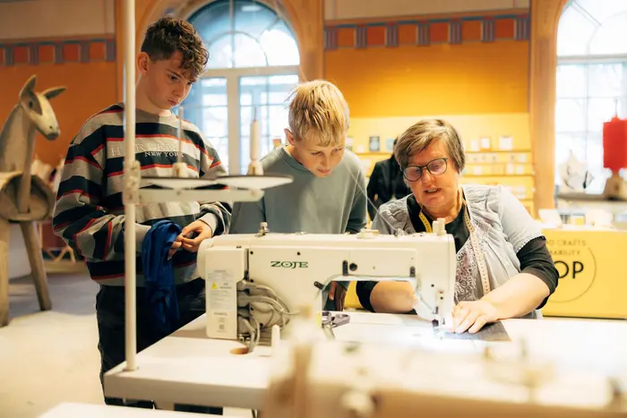 eine ältere Frau zeigt zwei Jungen etwas an einer Nähmaschine