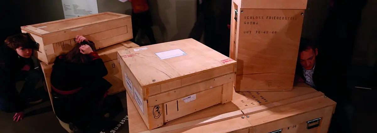 Menschen, die sich hinter Holzkisten setzen oder an diesen vorbeigehen. 